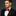 Man wearing a black suit with a red and black checkered bow tie and pocket square on a dark background