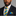 Man wearing a dark suit with a blue and yellow striped tie and pocket square on a gray background