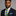 Man wearing a dark suit with a blue and yellow striped tie and pocket square on a gray background