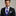 Man wearing a black suit with a purple plaid tie and blue pocket square on a gray background