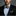 Man wearing a black suit with a checkered bow tie and pocket square against a gray background