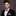 Man wearing a black suit with a red and white checkered bow tie and pocket square on a gray background