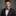 Man wearing a black suit with a purple checkered bow tie and pocket square on a gray background
