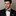 Man wearing a black suit with a patterned dark blue and beige plaid bow tie and pocket square against a neutral background
