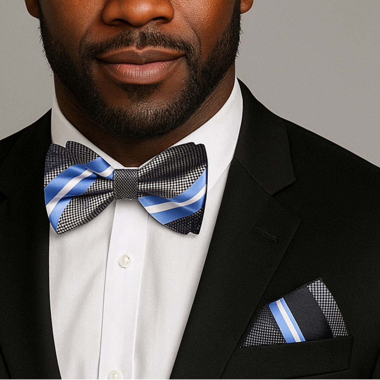 Man wearing a black suit with a blue, grey and white stripe bow tie and pocket square on a gray background