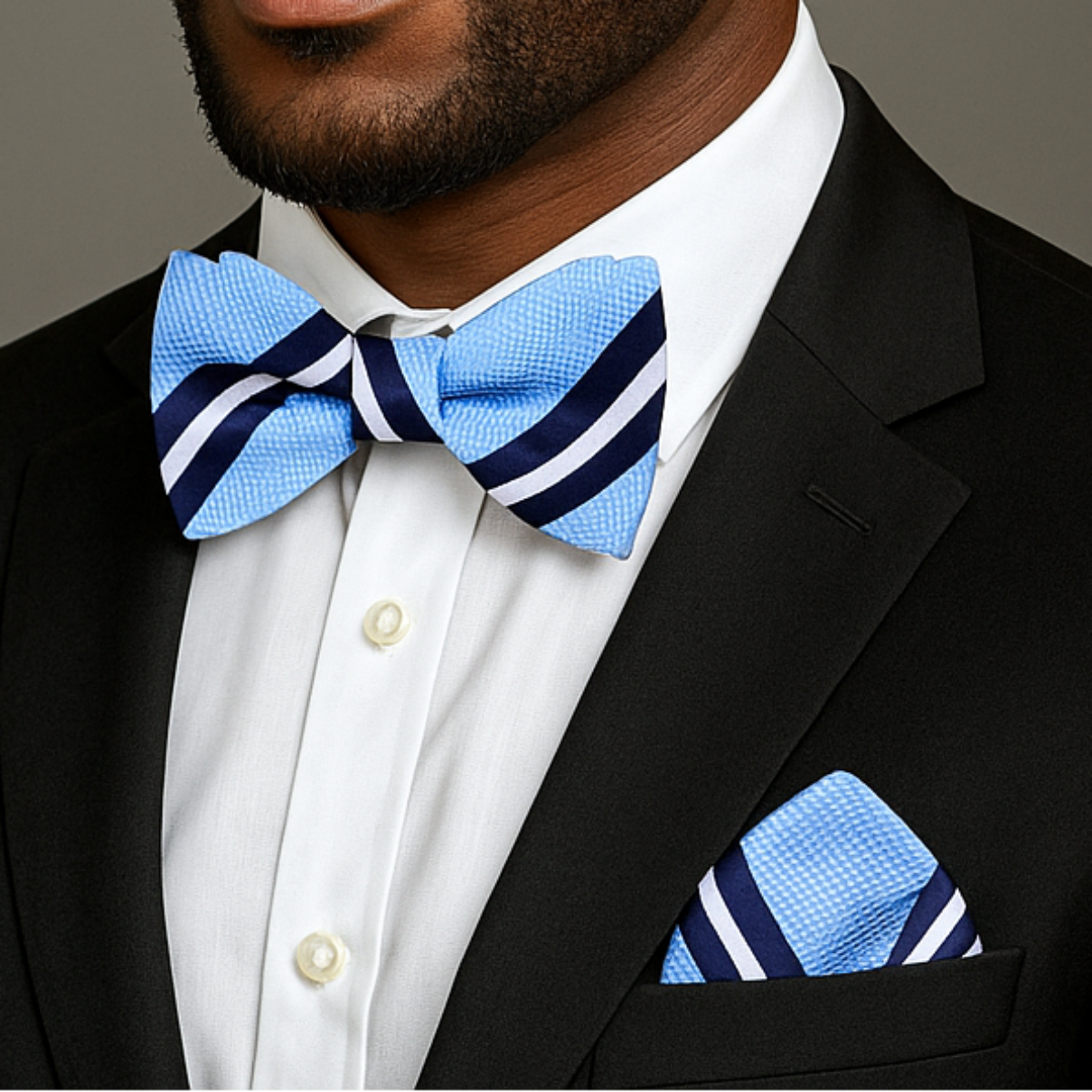 Man wearing a black suit with a blue stripe bow tie and pocket square on a gray background