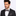 Man wearing a black suit with a patterned brown and grey check bow tie and pocket square on a light gray background