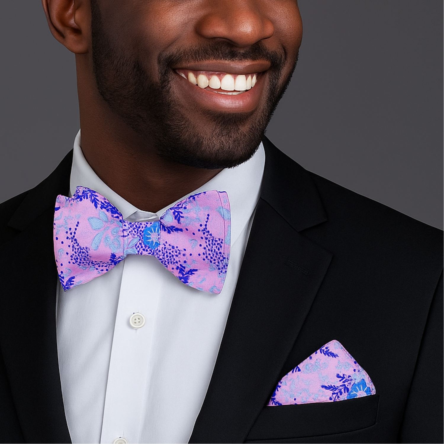 Man wearing a black suit with a colorful light purple and light blue floral bow tie and pocket square on a gray background