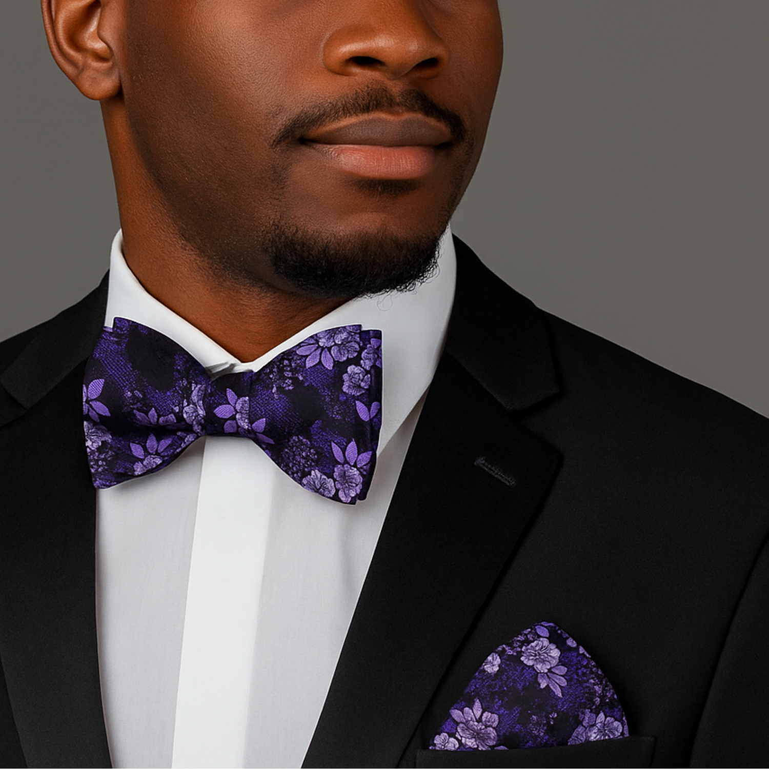 Man wearing a black suit with a purple floral bow tie and pocket square on a gray background