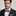 Man wearing a black suit with a white shirt and gray and black stripe bow tie against a plain background