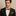 Man wearing a black suit with a patterned gold and red stripe bow tie and pocket square on a beige background