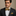 Man wearing a black suit with a gold stripe bow tie and pocket square against a beige background