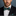 Man wearing a black suit with a green and brown striped bow tie and pocket square against a gray background