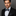 Man wearing a black suit with a green an blue plaid bow tie and pocket square on a gray background