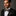 Man wearing a black suit with a light yellow blue and black plaid bow tie and pocket square on a gray background