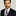 Man wearing a black suit with a striped plum and white stripe tie and red pocket square on a gray background