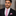 Man wearing a black suit with a thin pink tie with blue dots and pocket square against a blurred background
