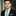 Man wearing a black suit with a green with blue dots thin tie and blue pocket square, standing against a blurred background.