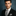 Man wearing a black suit with a checkered black and light grey tie and pocket square against a dark background