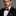 Man wearing a black suit with a white shirt and a pink paisley bow tie and pocket square on a gray background