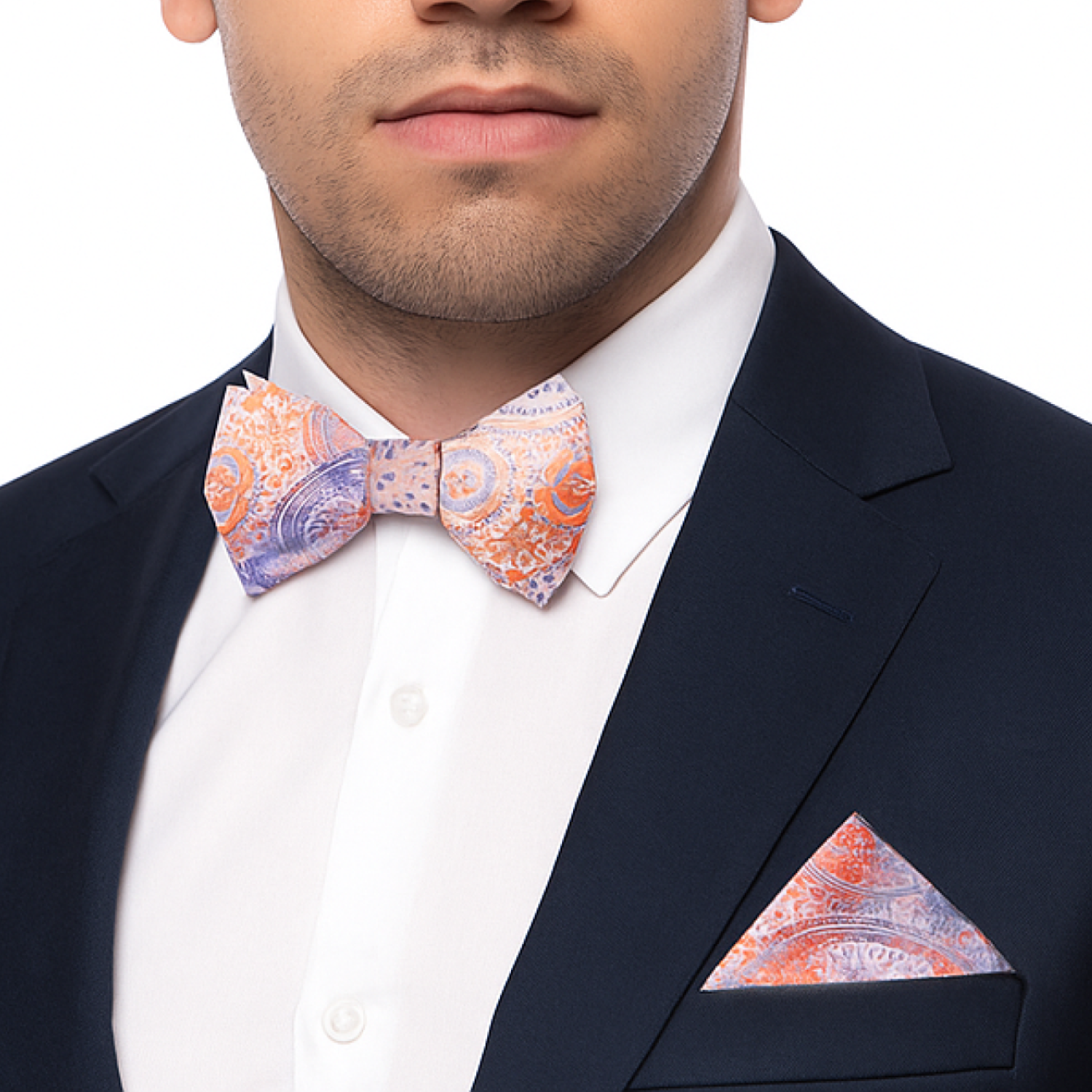 Man wearing a navy suit with a peach and purple paisley patterned bow tie and pocket square on a white background