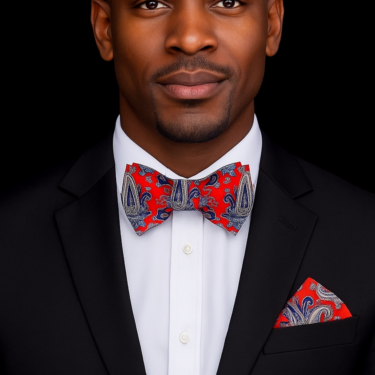 Man wearing a black suit with a red and blue paisley bow tie and pocket square on a black background