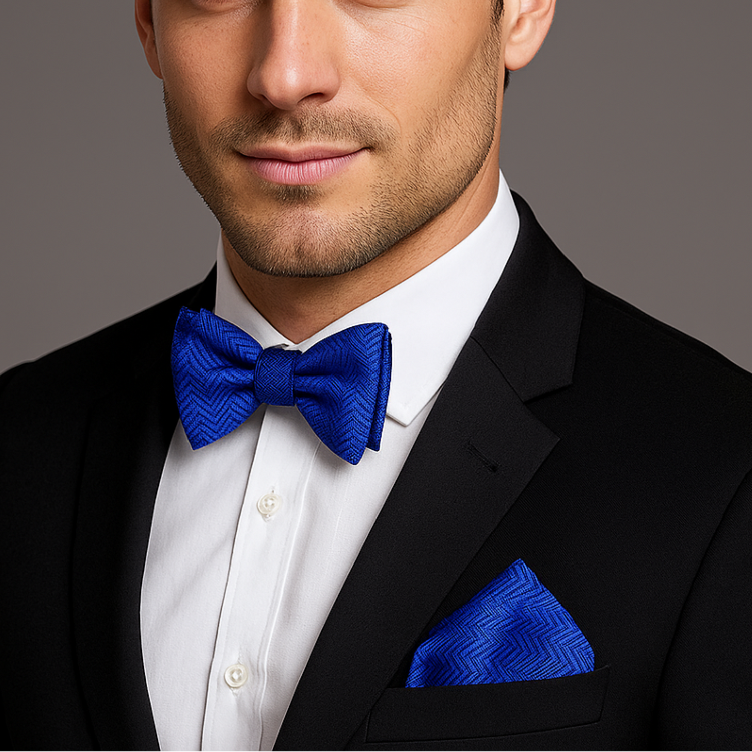 Man wearing a black suit with a white shirt and blue bow tie and pocket square on a gray background