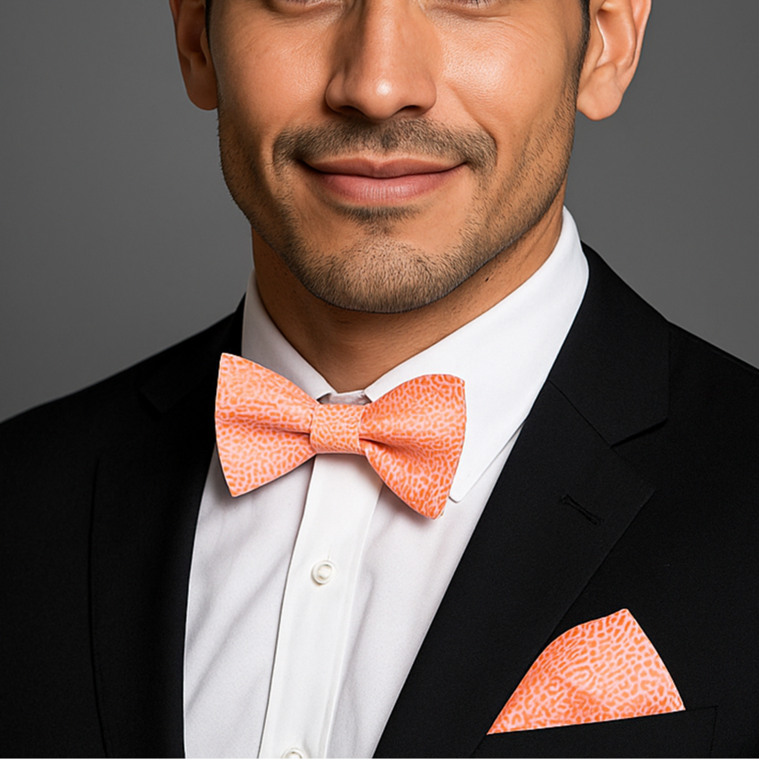 Man wearing a black suit with a coral bow tie and pocket square on a gray background