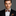 Man wearing a black suit with a colorful orange, yellow, red and black swirls bow tie and matching pocket square on a gray background