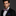 Man wearing a black suit with a colorful pink, blue paisley bow tie and accenting blue and pink pocket square on a dark background