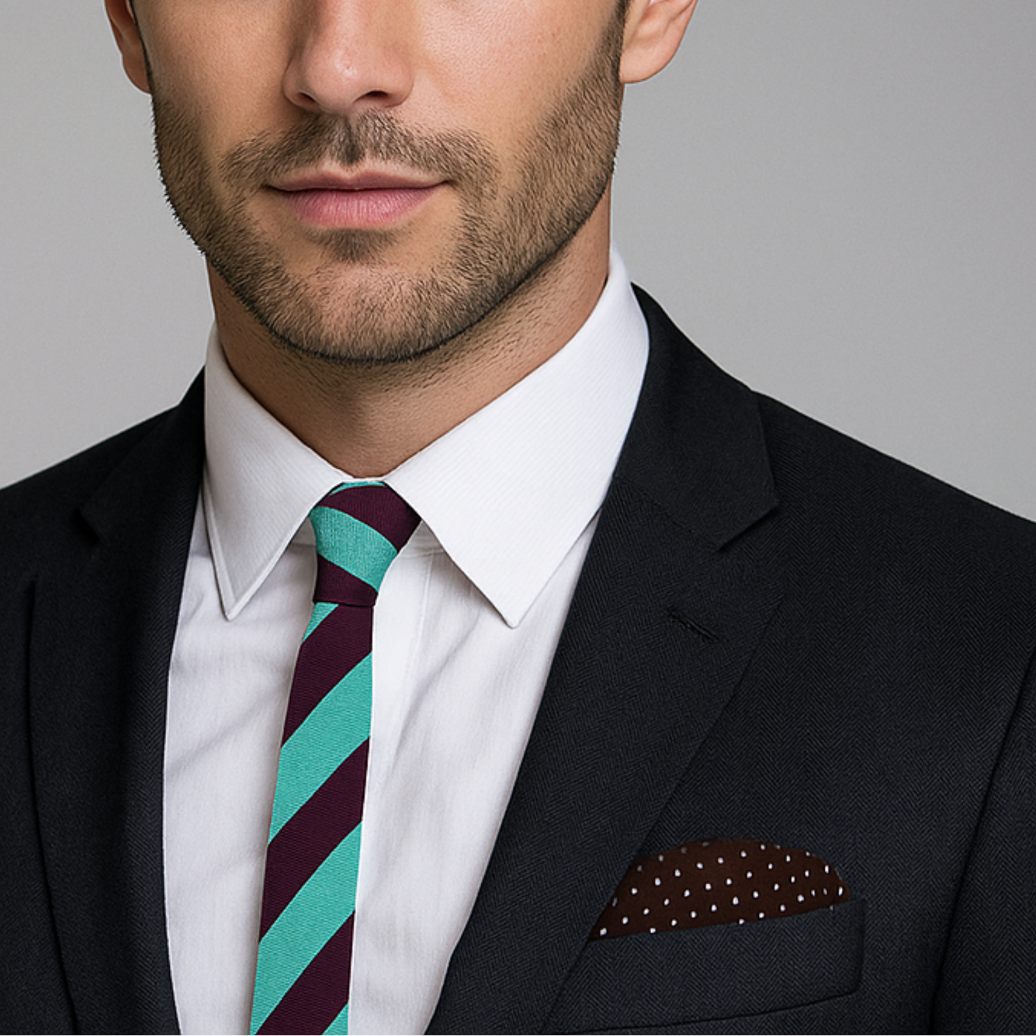 Man wearing a black suit with a brown and green striped thin tie and accenting brown and white polka square against a gray background