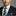 Man wearing a black suit with a brown and green striped thin tie and a square on a gray background