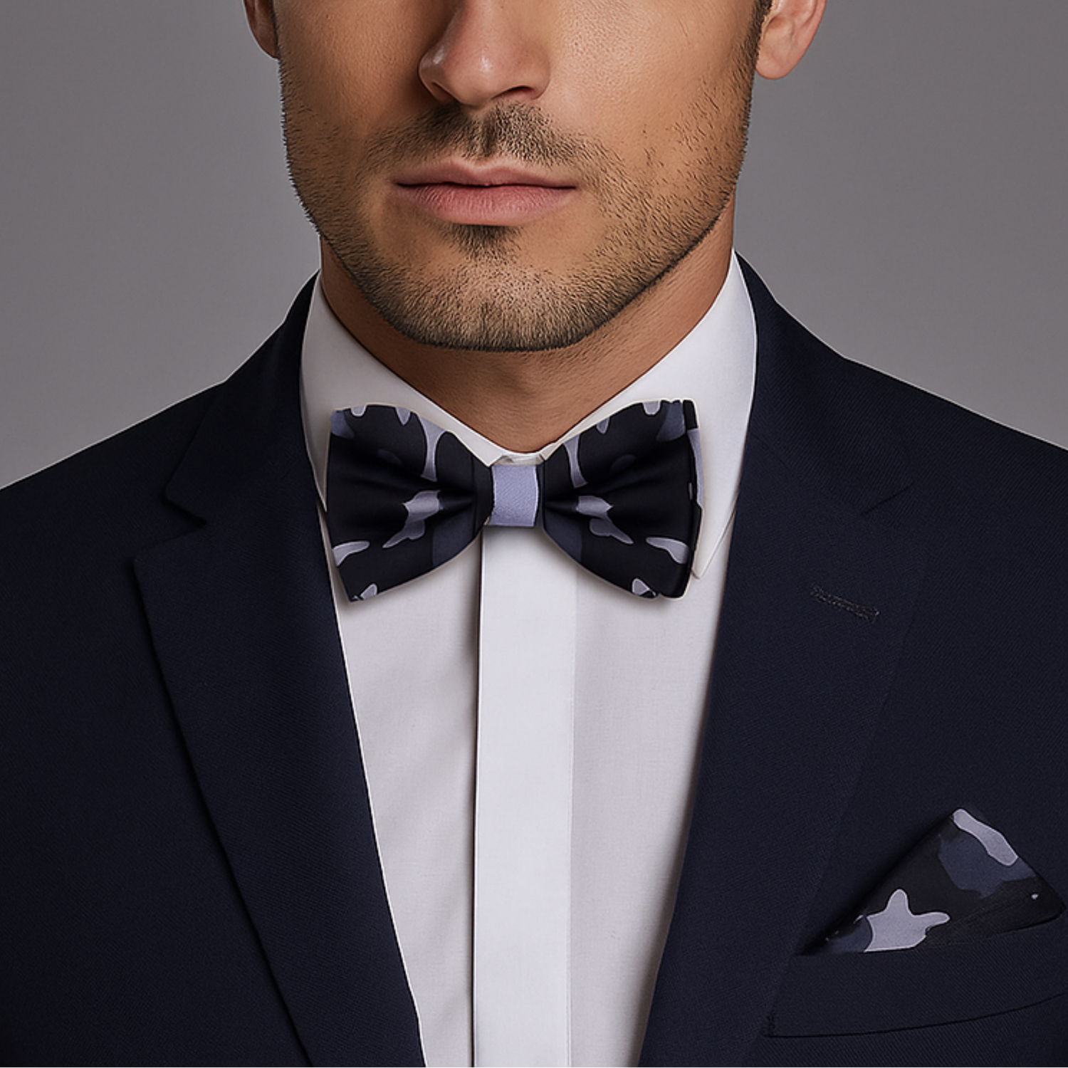 Man wearing a dark suit with a A Black, Grey Camo Fleck Pattern Self Tie Bow Tie, Matching Pocket Square against a gray background