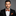 Man wearing a black suit with an orange and black patterned bow tie and pocket square on a gray background