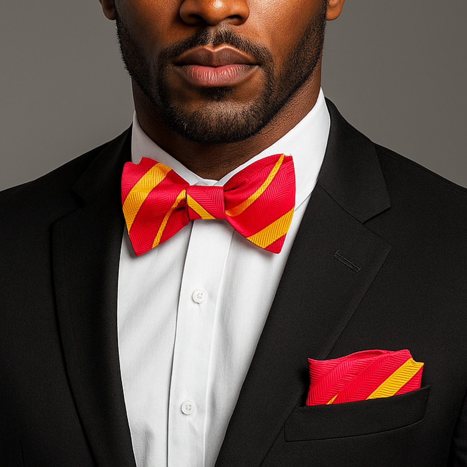 Man wearing a black suit with a red and yellow bow tie and pocket square against a gray background