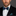 Man wearing a black suit with a green and blue bow tie and pocket square on a gray background