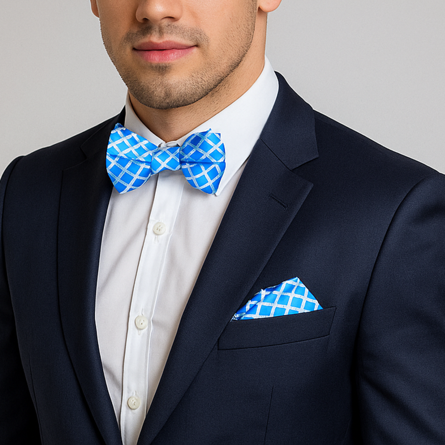 Man wearing a navy suit with a white shirt, blue patterned bow tie, and pocket square against a gray background.