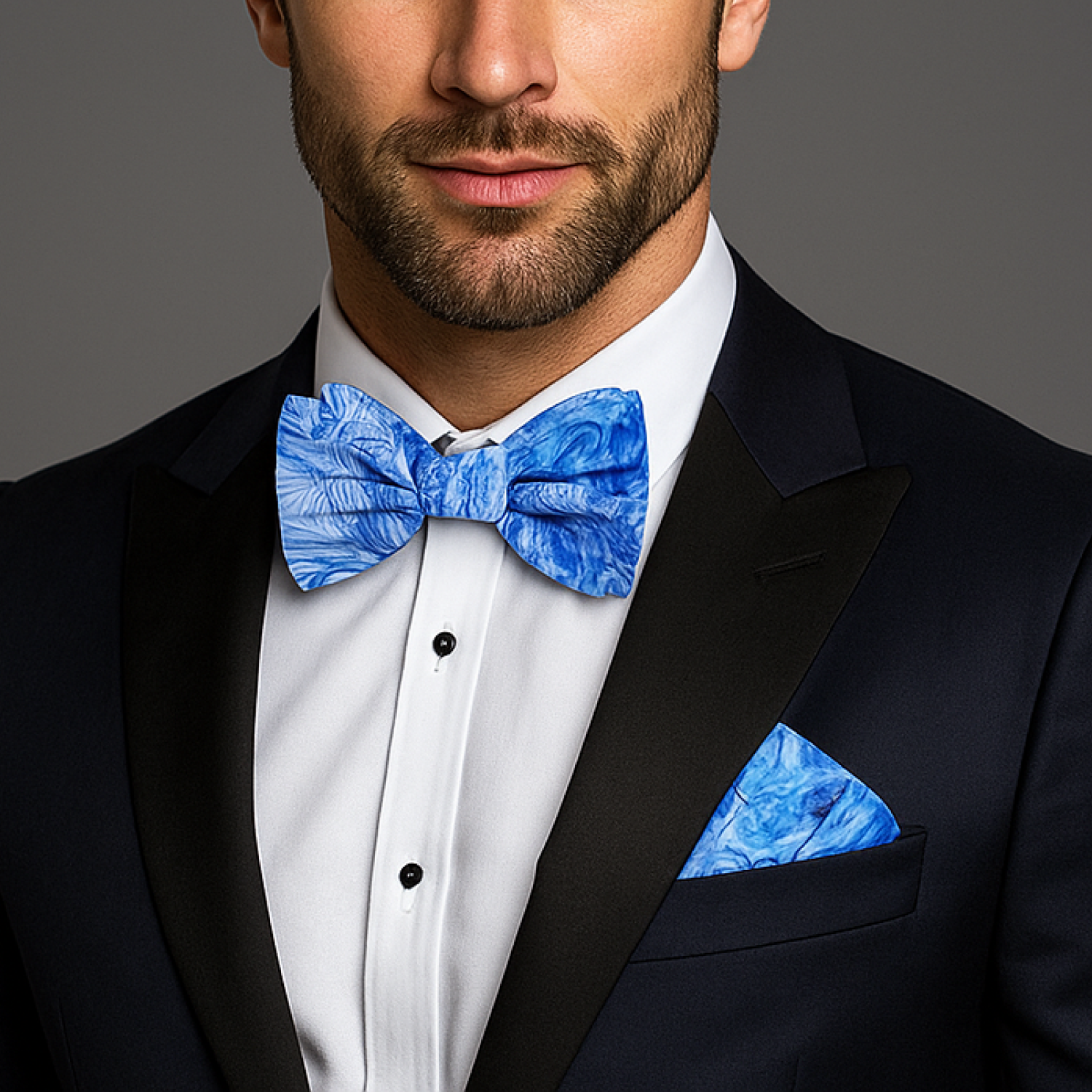 Man wearing a black tuxedo with a blue bow tie and pocket square against a gray background