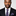 Man wearing a black suit with a patterned tie and pocket square on a plain background