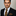 Man wearing a black suit with a checkered tie and pocket square against a neutral background