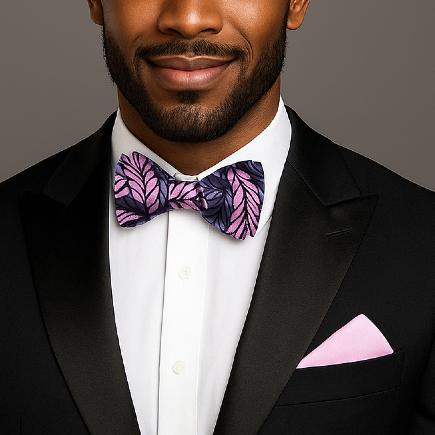 Man wearing a black tuxedo with a grey and pink bow tie and pocket square on a gray background