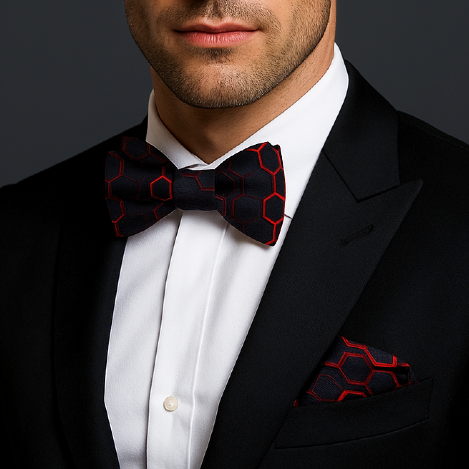 Man wearing a black tuxedo with a red geometric patterned bow tie and pocket square on a dark background.