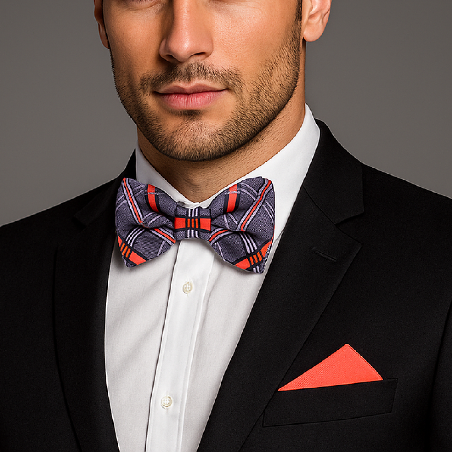 Man wearing a black suit with a grey and coral plaid bow tie and red pocket square on a gray background