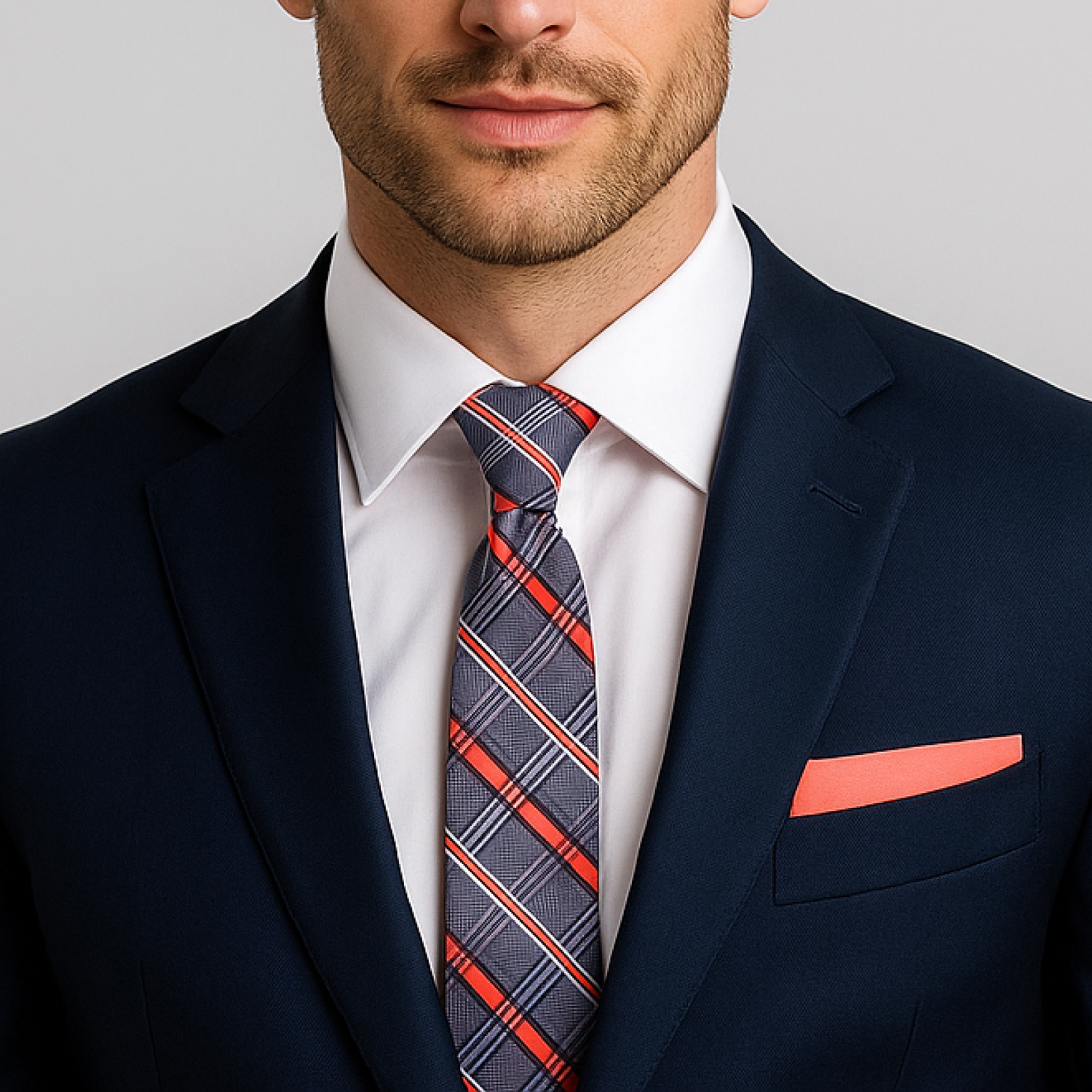 Man wearing a navy suit with a plaid tie and coral pocket square on a gray background