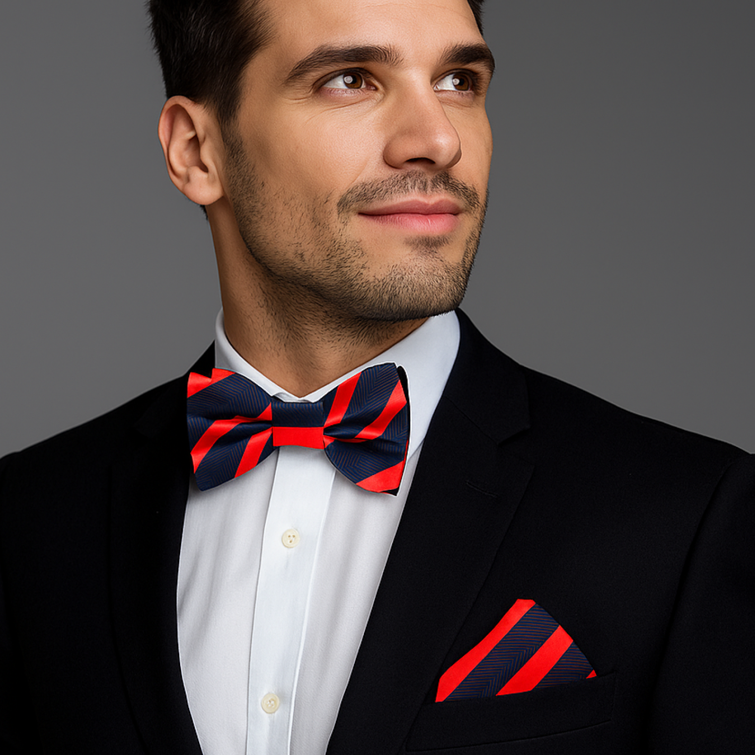 Man wearing a black suit with a red and blue striped bow tie and pocket square on a gray background
