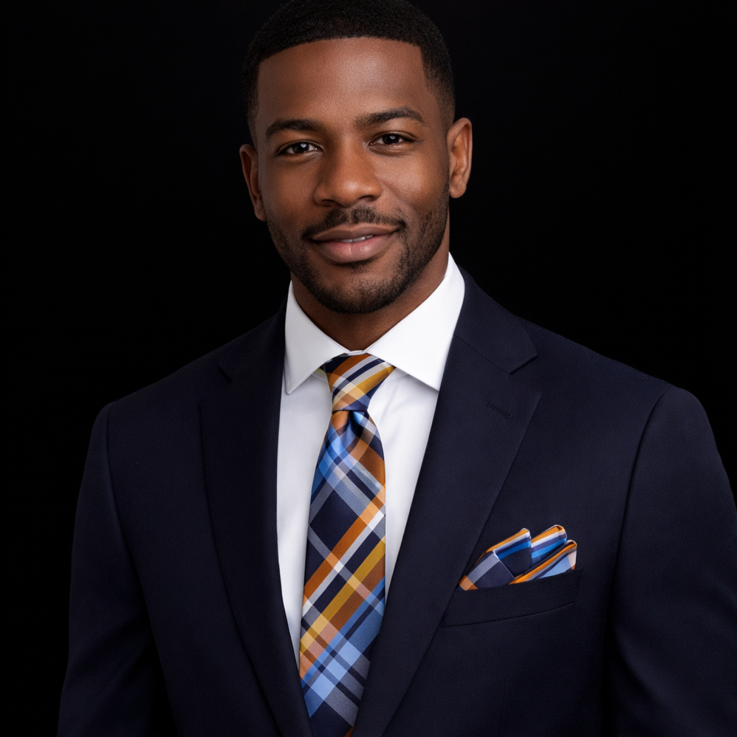 Man wearing a navy suit with a plaid tie and pocket square on a black background