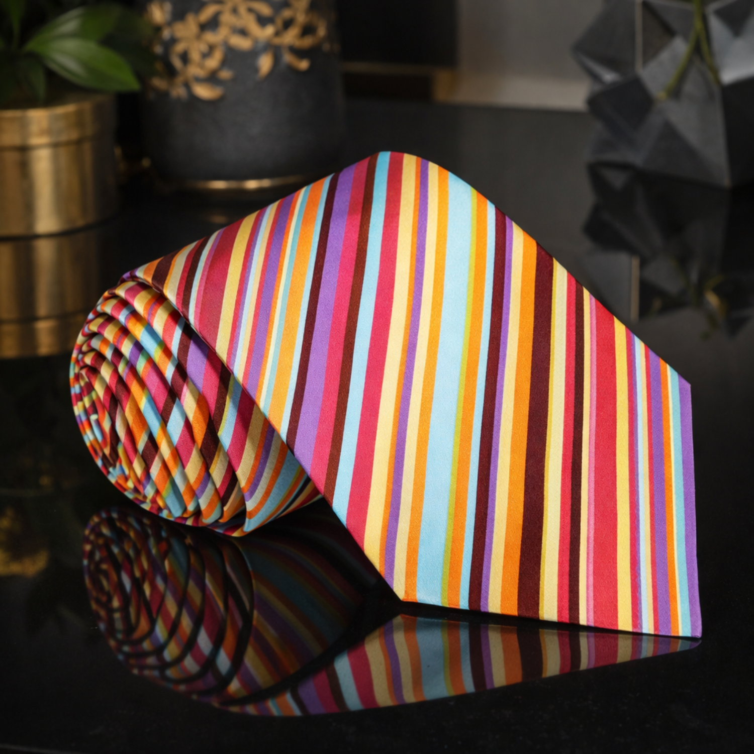 Roll of colorful striped tie on a reflective black surface with decorative items in the background.