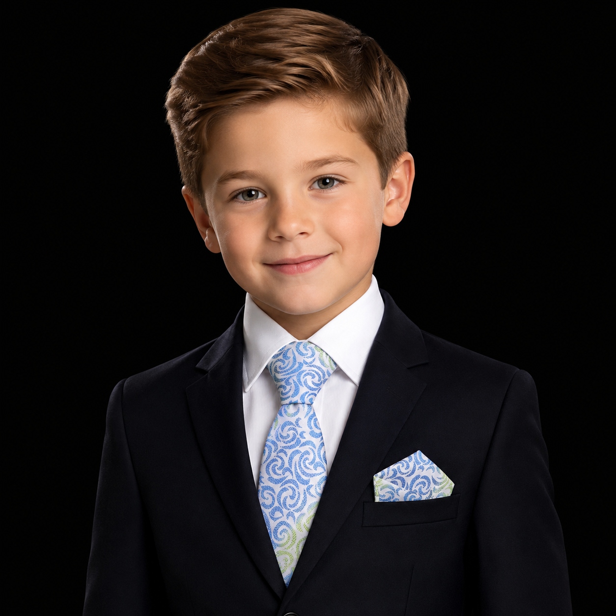A young boy with brown hair wearing a dark navy suit, white dress shirt, and a light blue and white patterned tie with a matching pocket square, set against a solid black background.