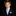 A young boy with brown hair wearing a dark navy suit, white dress shirt, and a light blue and white patterned tie with a matching pocket square, set against a solid black background.