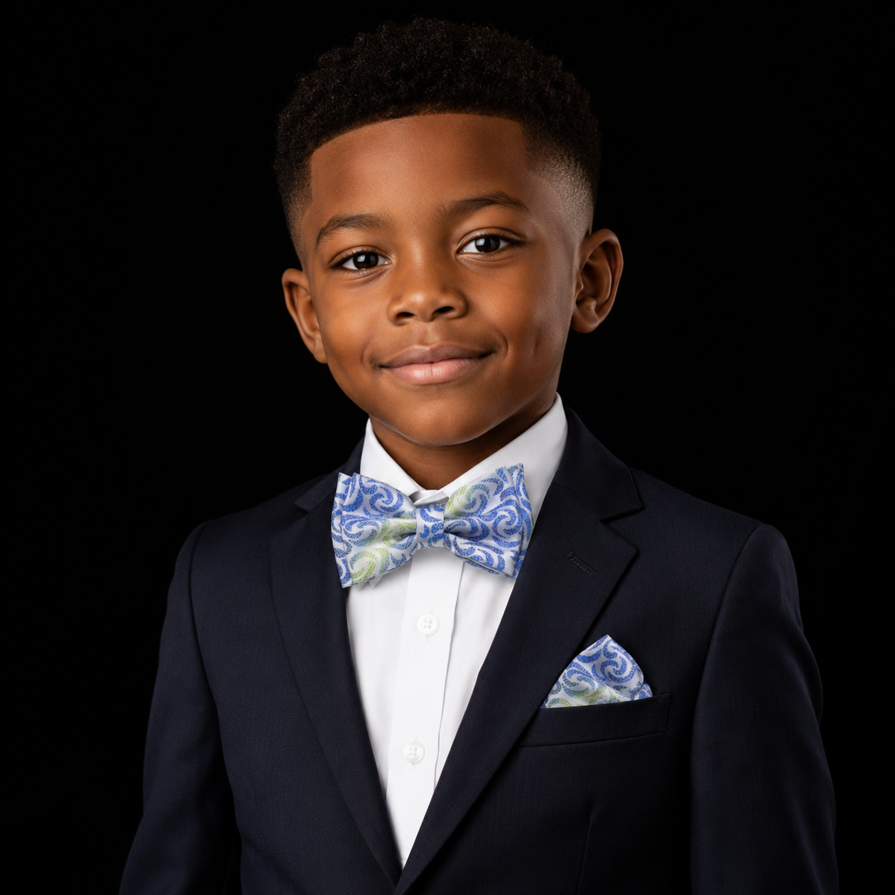 A young boy wearing a navy blue suit jacket, white dress shirt, and a blue and green paisley bow tie with a matching pocket square, set against a solid black background.
